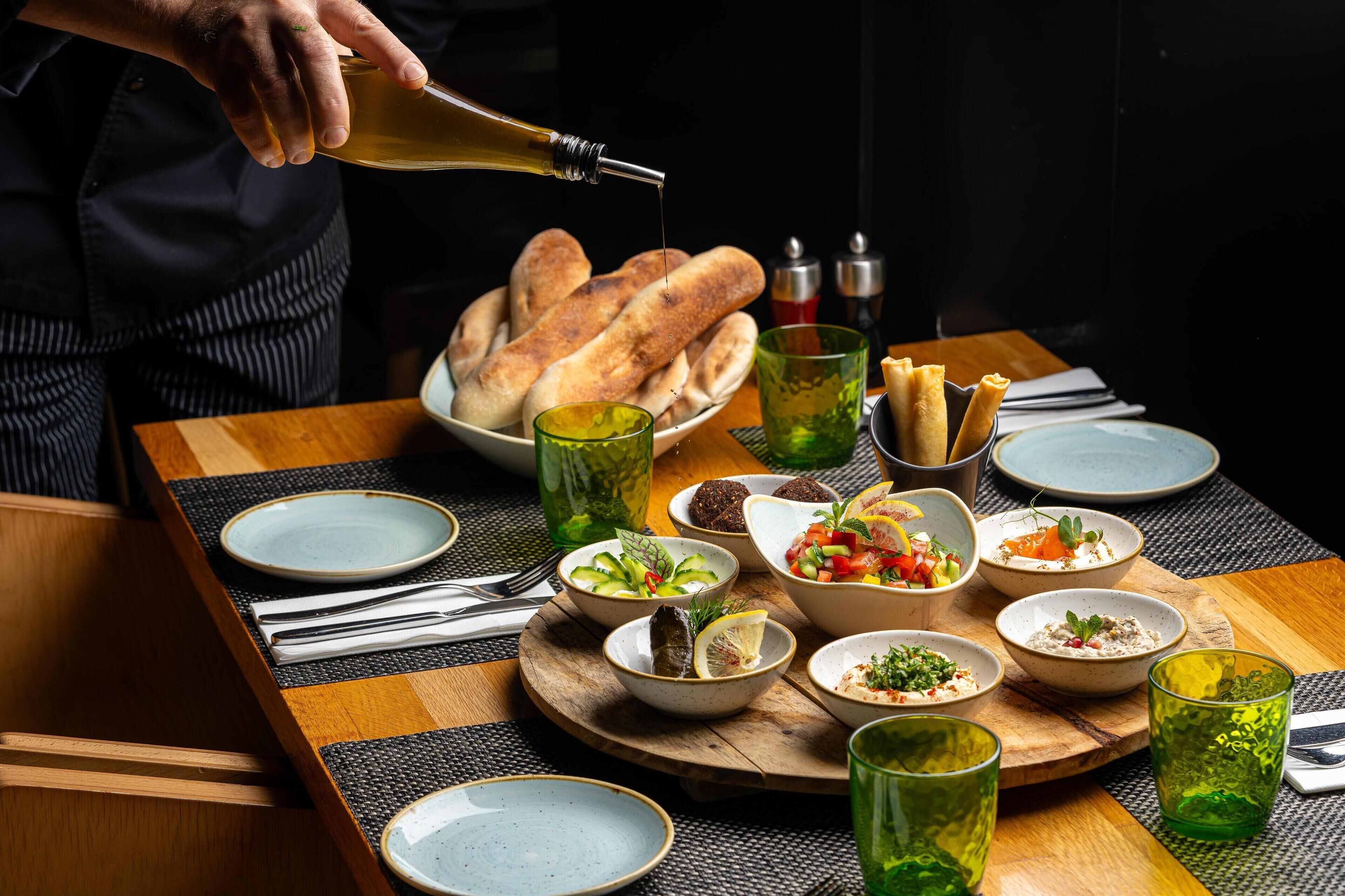 A table set with various dishes, including bread, salads, and bowls, with a person pouring oil.