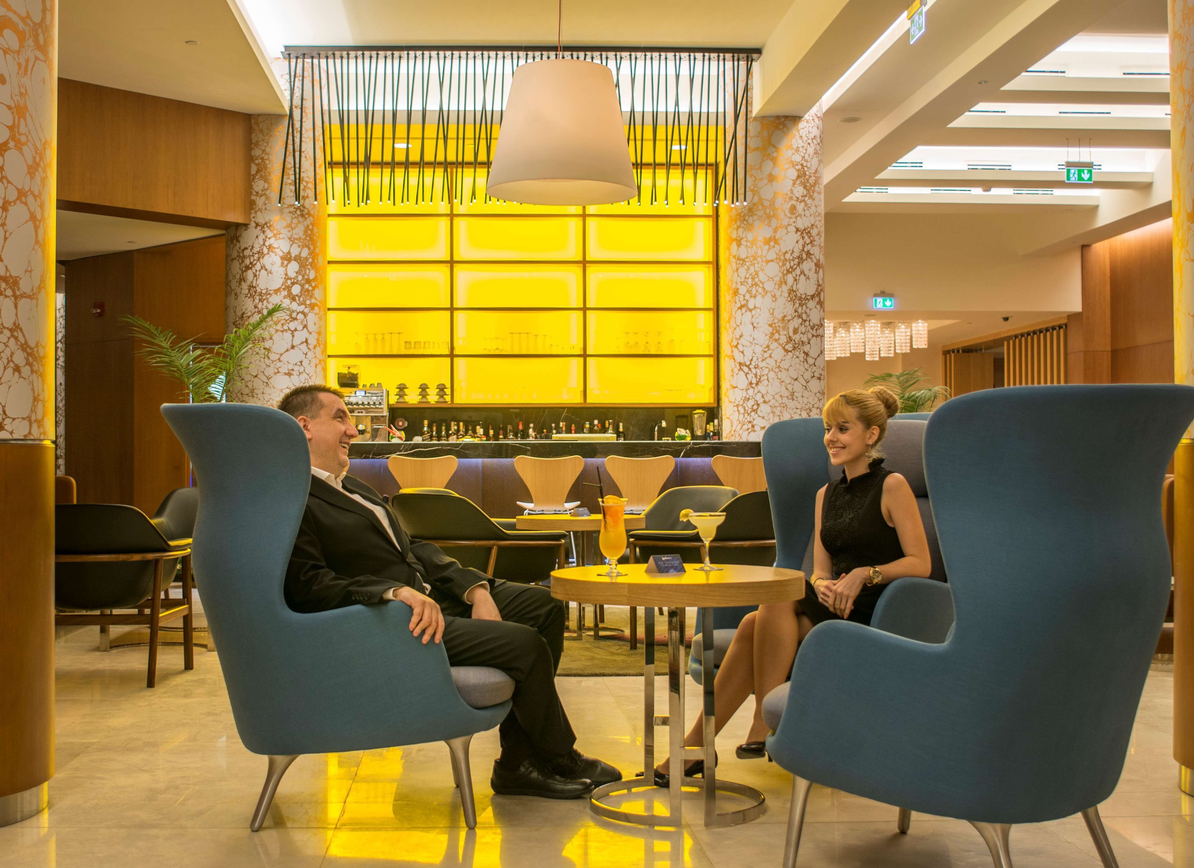 A modern lounge area with blue chairs, a central table with drinks, and a yellow-lit background.