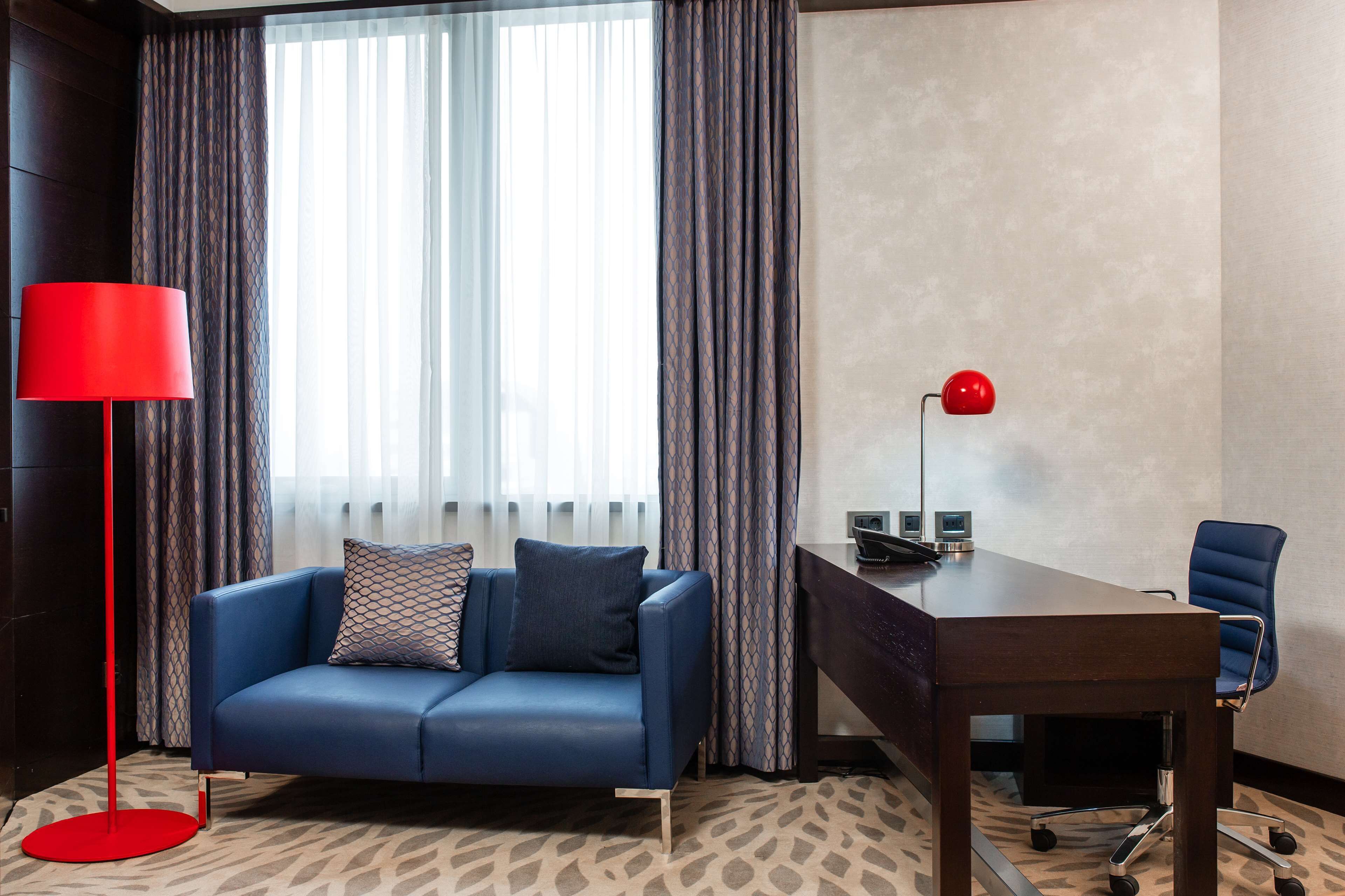 A modern hotel room with a blue sofa, a red floor lamp, a wooden desk, and a blue office chair.