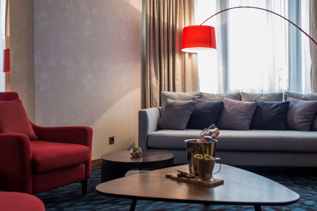 Une chambre d’hôtel moderne avec un fauteuil rouge, un canapé gris, une table basse ronde sur laquelle se trouve un panier de fruits, et un lampadaire rouge.
