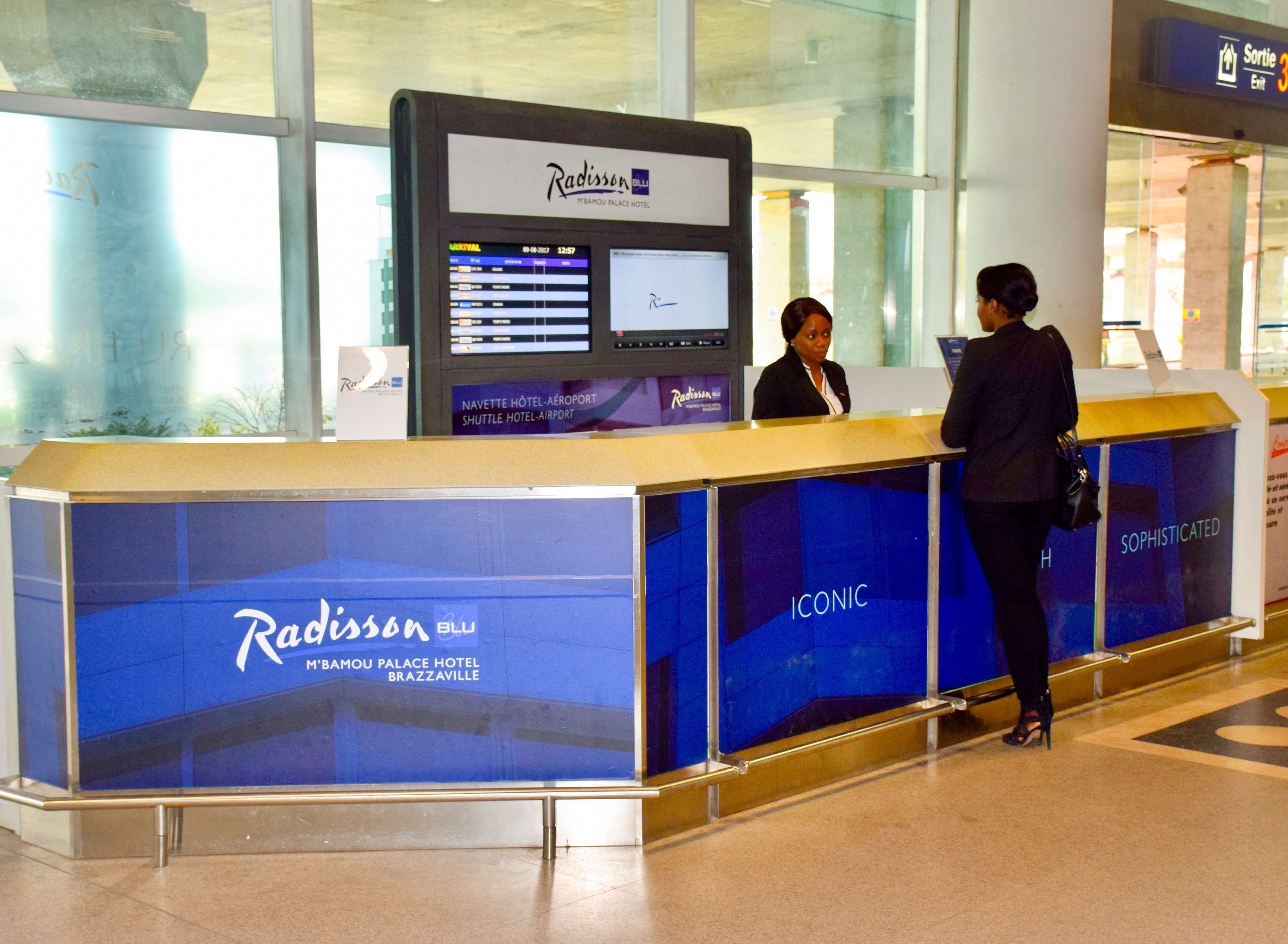 Le comptoir de réception d'un hôtel Radisson Blu, doté d'un écran numérique et d'une enseigne indiquant « M'BAMOU PALACE HOTEL ».