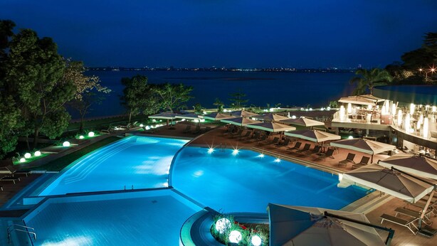 A large, illuminated swimming pool with lounge chairs and umbrellas, surrounded by trees and a view of the sea at night. - Radisson Blu M'Bamou Palace Hotel (Brazzaville)