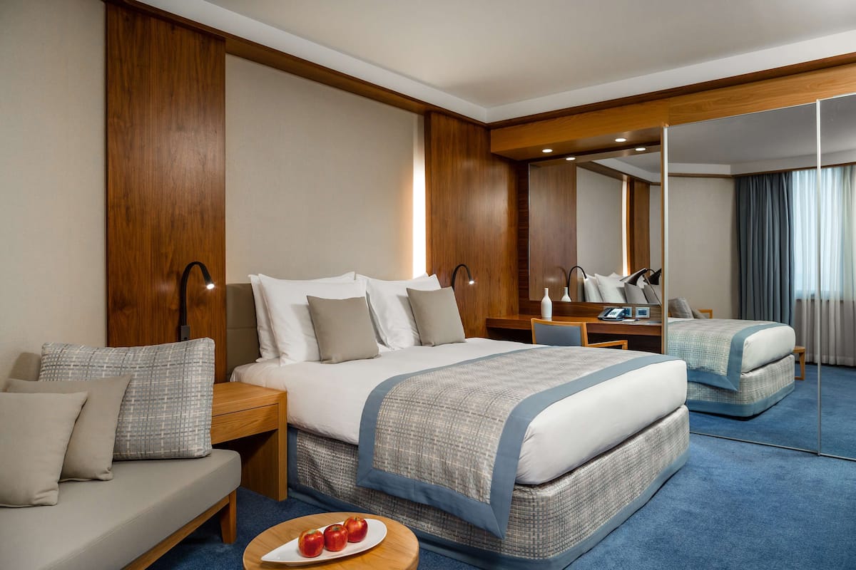 Une chambre d’hôtel avec un grand lit, un canapé, une table ronde avec des pommes et un miroir qui reflète la pièce.