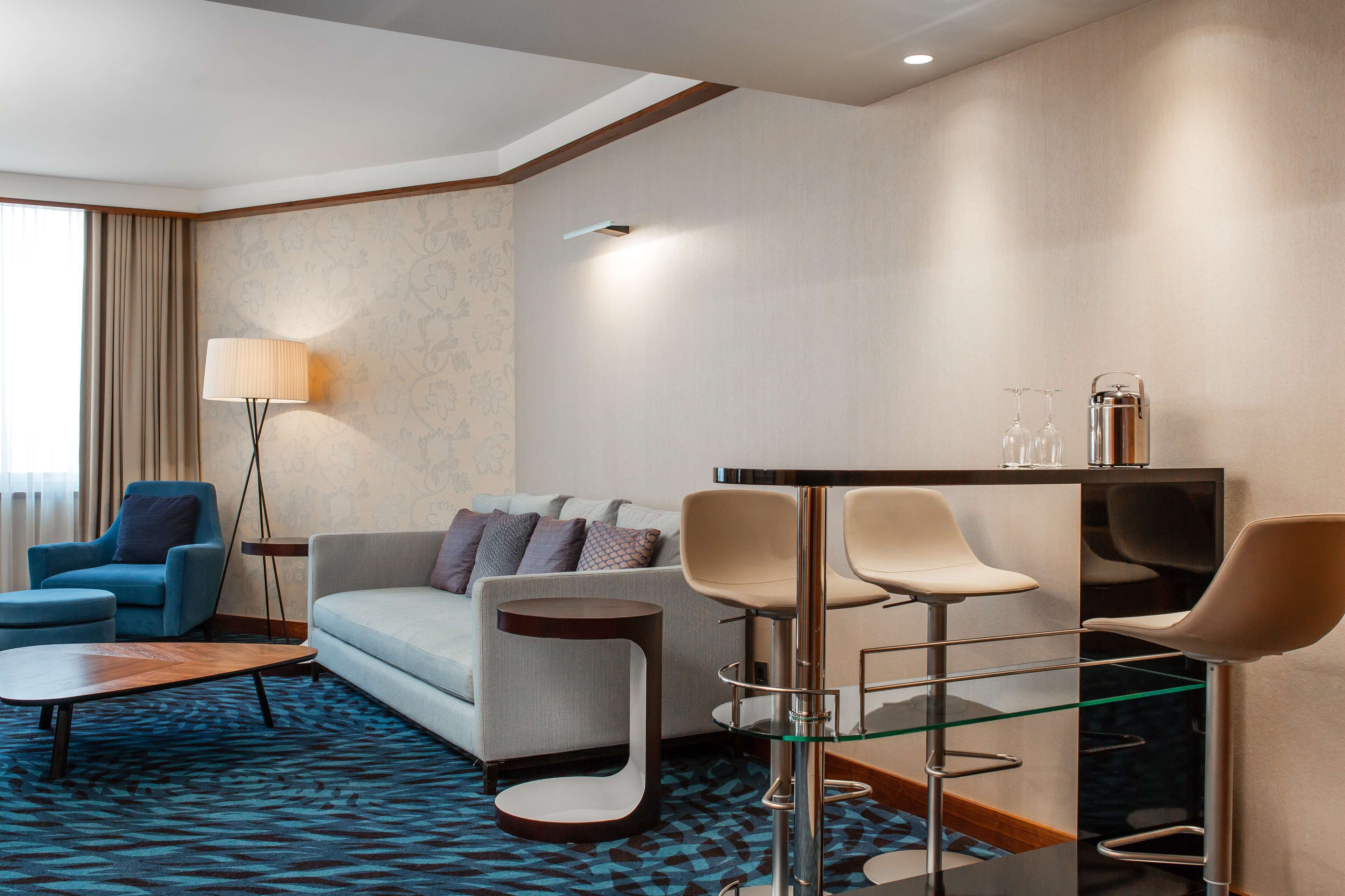 A modern hotel room with a sofa, armchair, and a bar area with stools.
