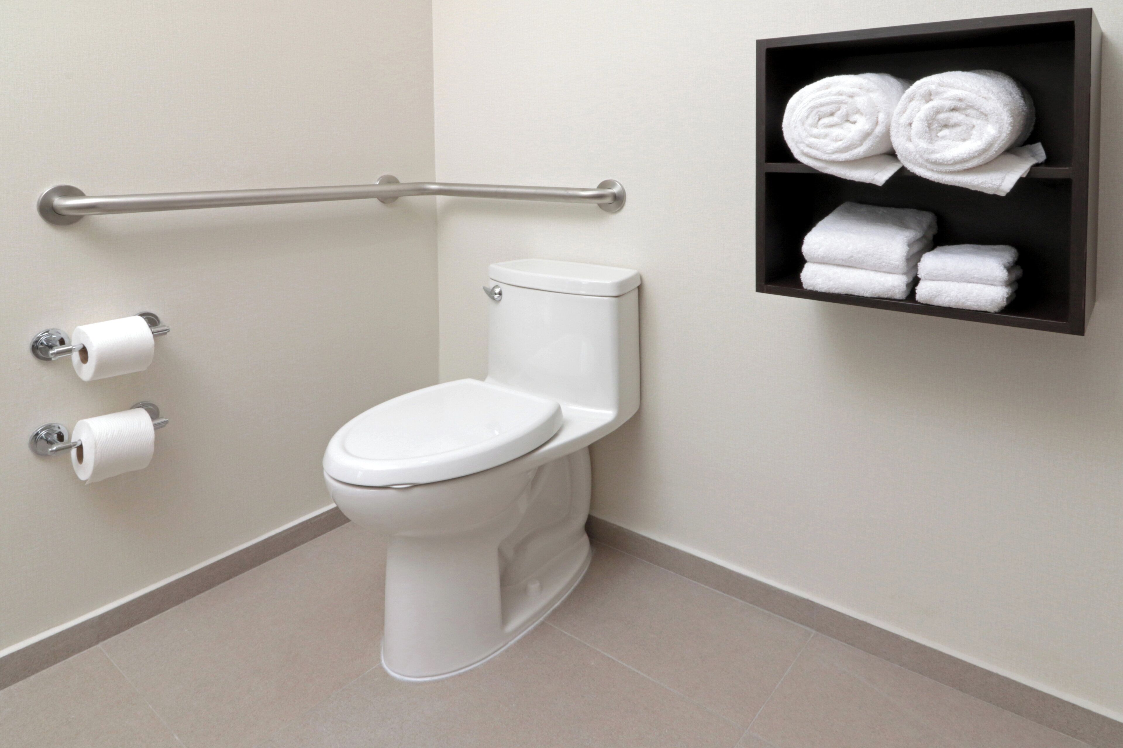 A toilet with a grab bar, two rolls of toilet paper, and a shelf with towels.