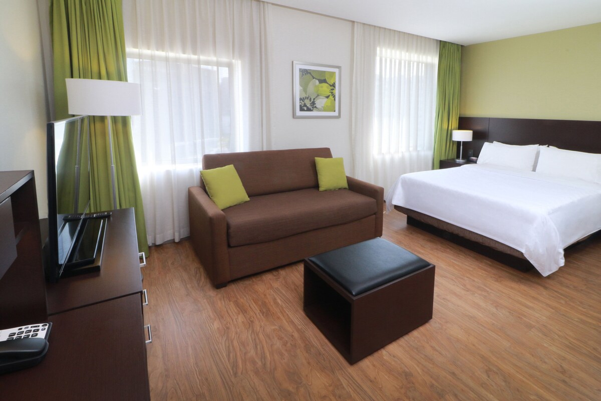 Una habitación de hotel con una cama, un sofá marrón con cojines verdes, una mesita y un escritorio con lámpara.