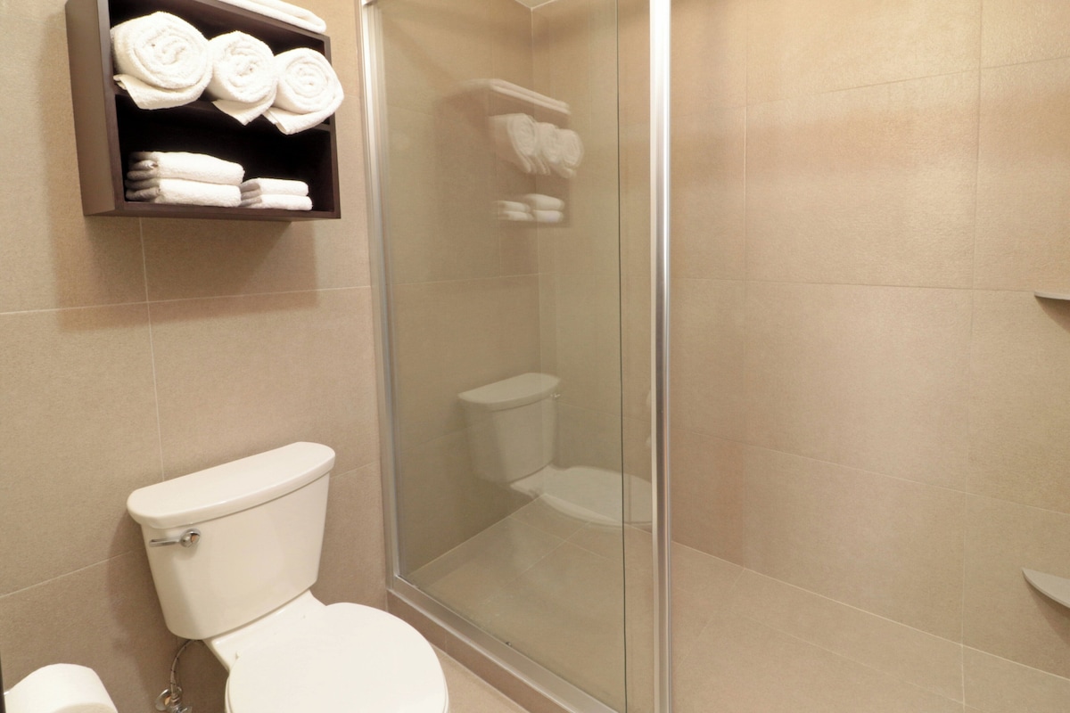 Un baño con inodoro, ducha con mampara de vidrio y estante con toallas enrolladas.