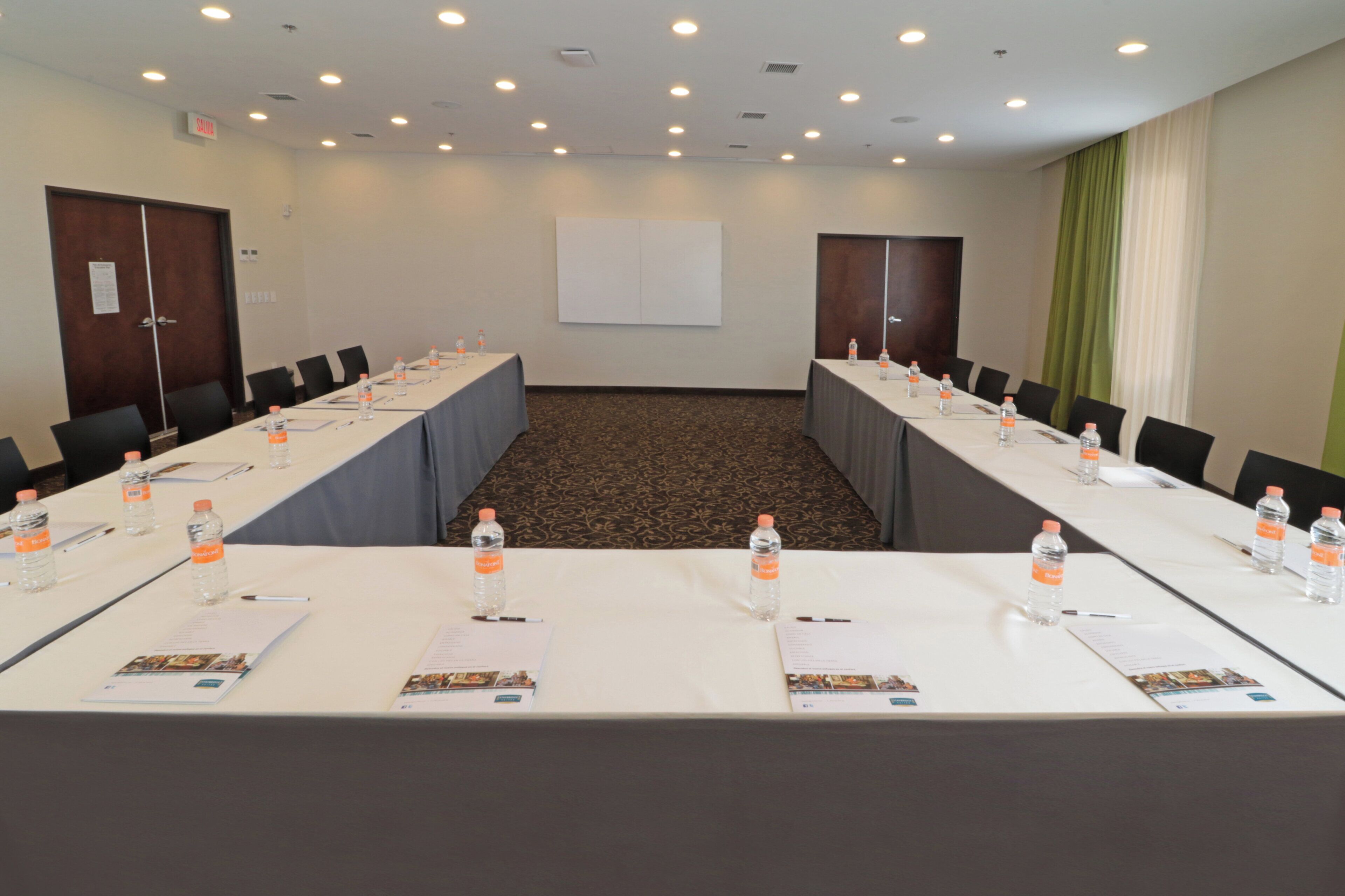 Un salón de conferencias con una larga mesa, sillas, botellas de agua y una pantalla de proyector.