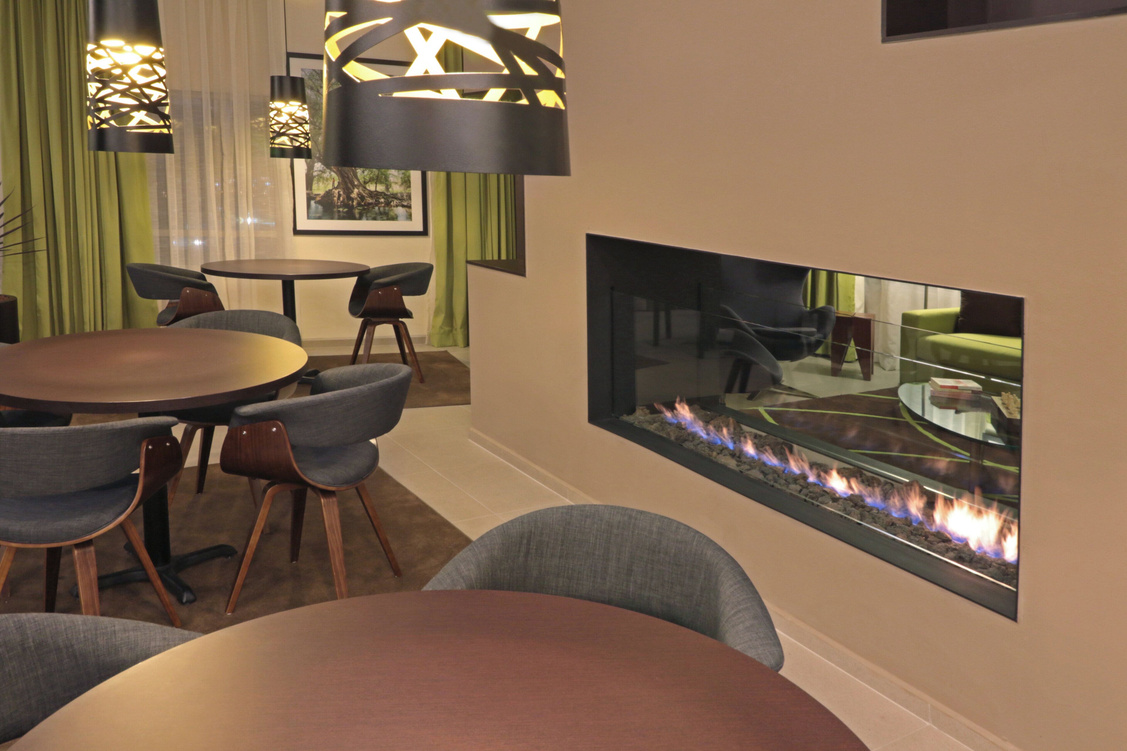 A modern interior with a round dining table, grey upholstered chairs, a glass-top coffee table, and a built-in fireplace with visible flames.