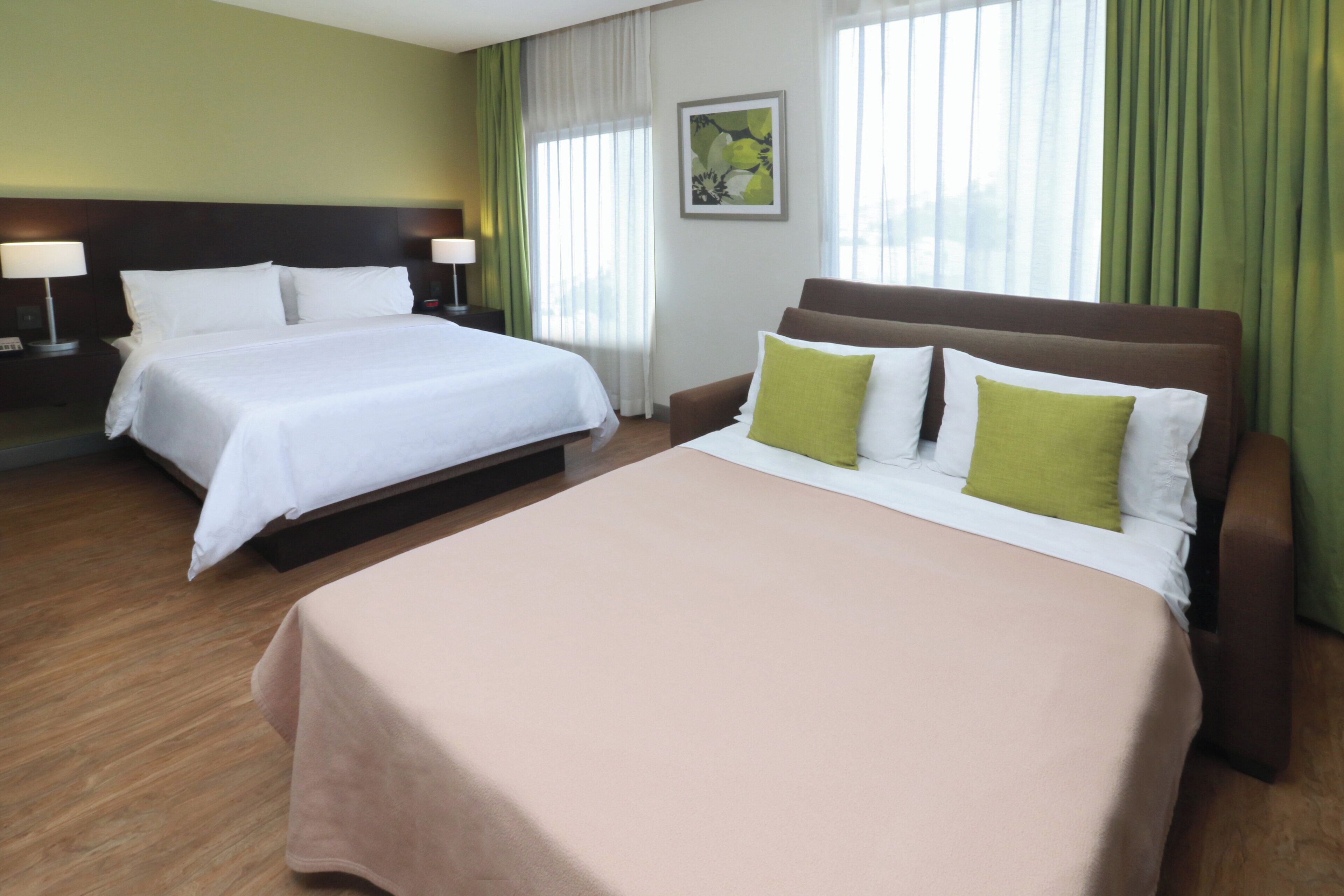 A hotel room with two beds, a large bed in the center and a smaller one on the left. There is a nightstand with a lamp on each side of the smaller bed. The room has a wooden floor and green curtains on the windows.