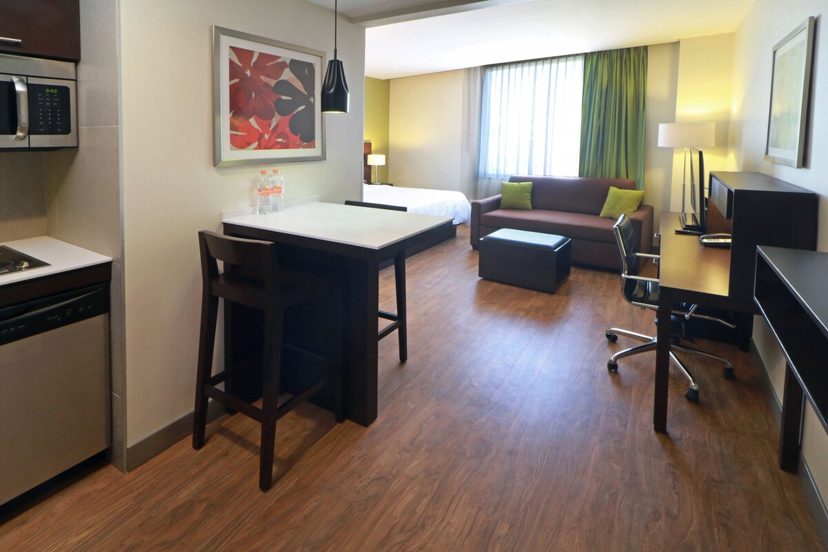 Habitación de hotel con kitchenette, zona de comedor, sofá, televisor y ventana con cortinas.