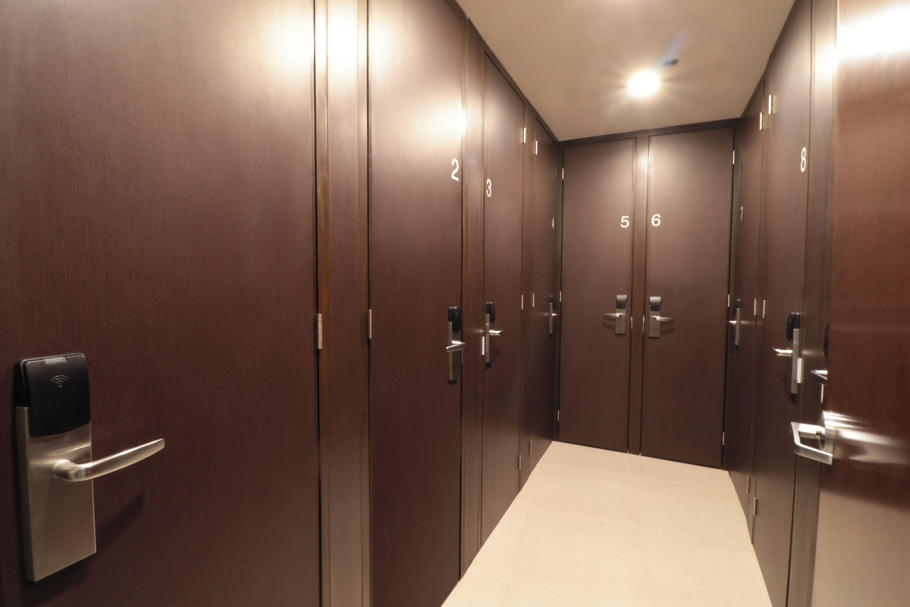 Un couloir avec des portes en bois numérotées de 1 à 8, un distributeur de gel hydroalcoolique et un luminaire au plafond.