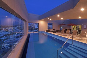 An indoor swimming pool with clear glass walls, a view of a cityscape, and a seating area with lounge chairs and umbrellas. - Staybridge Suites San Luis Potosi by IHG (San Luis Potosi)