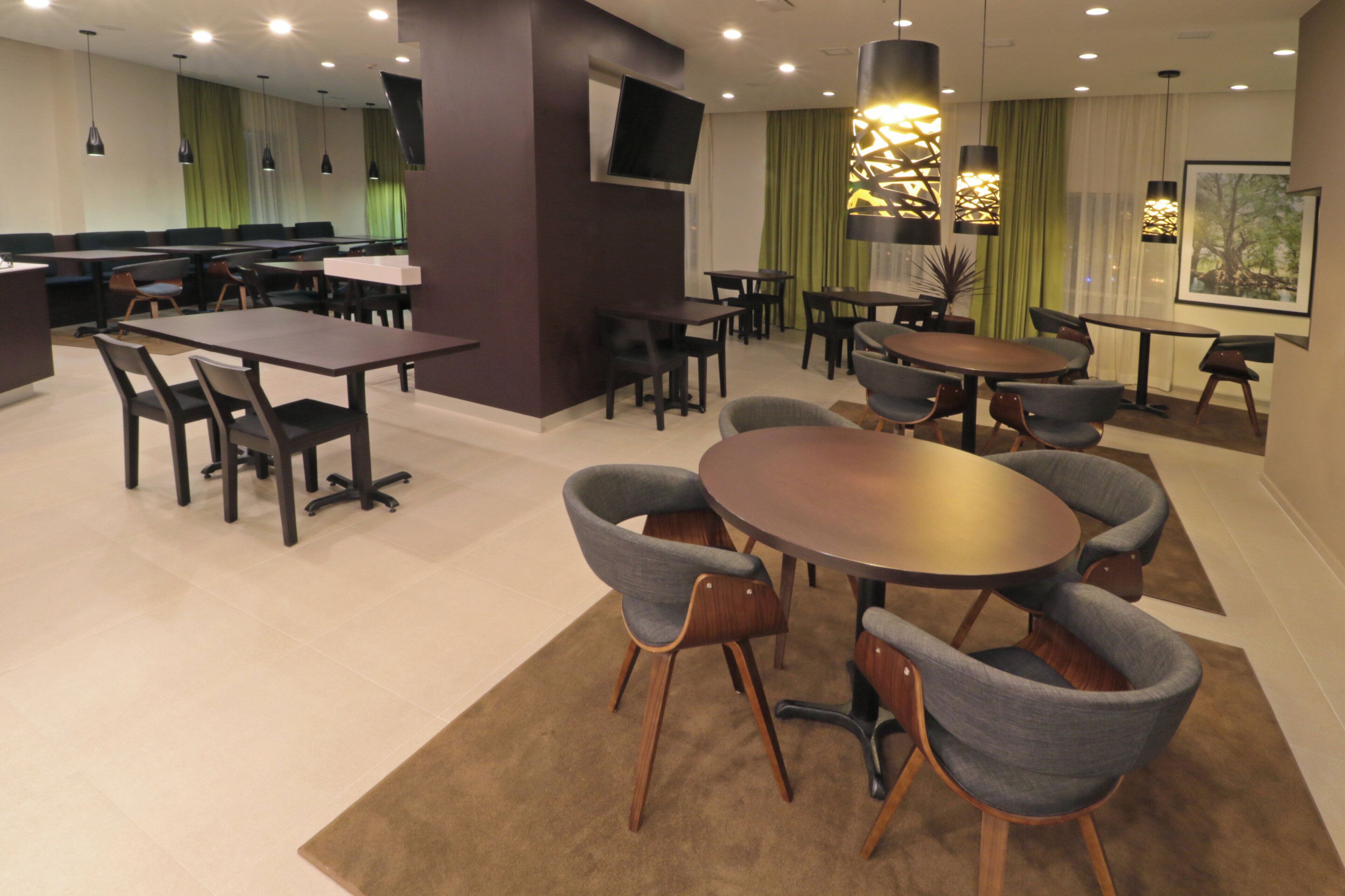 A modern dining area with multiple tables and chairs, a flat-screen TV mounted on the wall, and a large window with curtains.