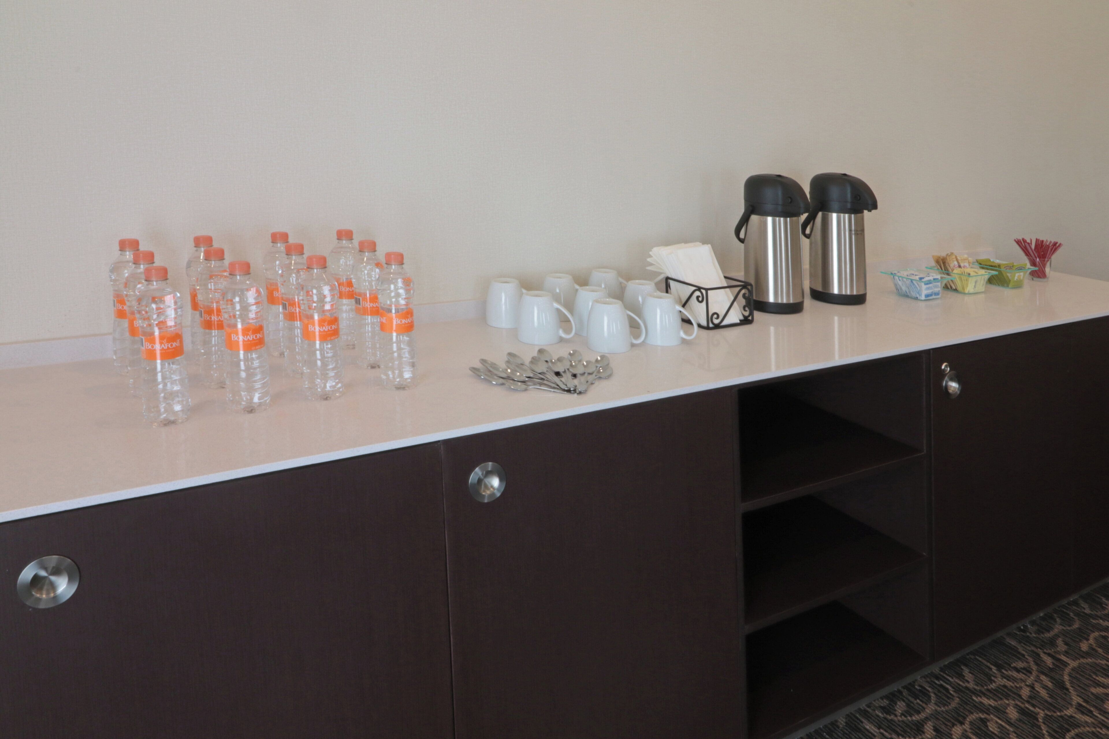 A hotel room counter with water bottles, white mugs, a coffee maker, and a small bowl of snacks.