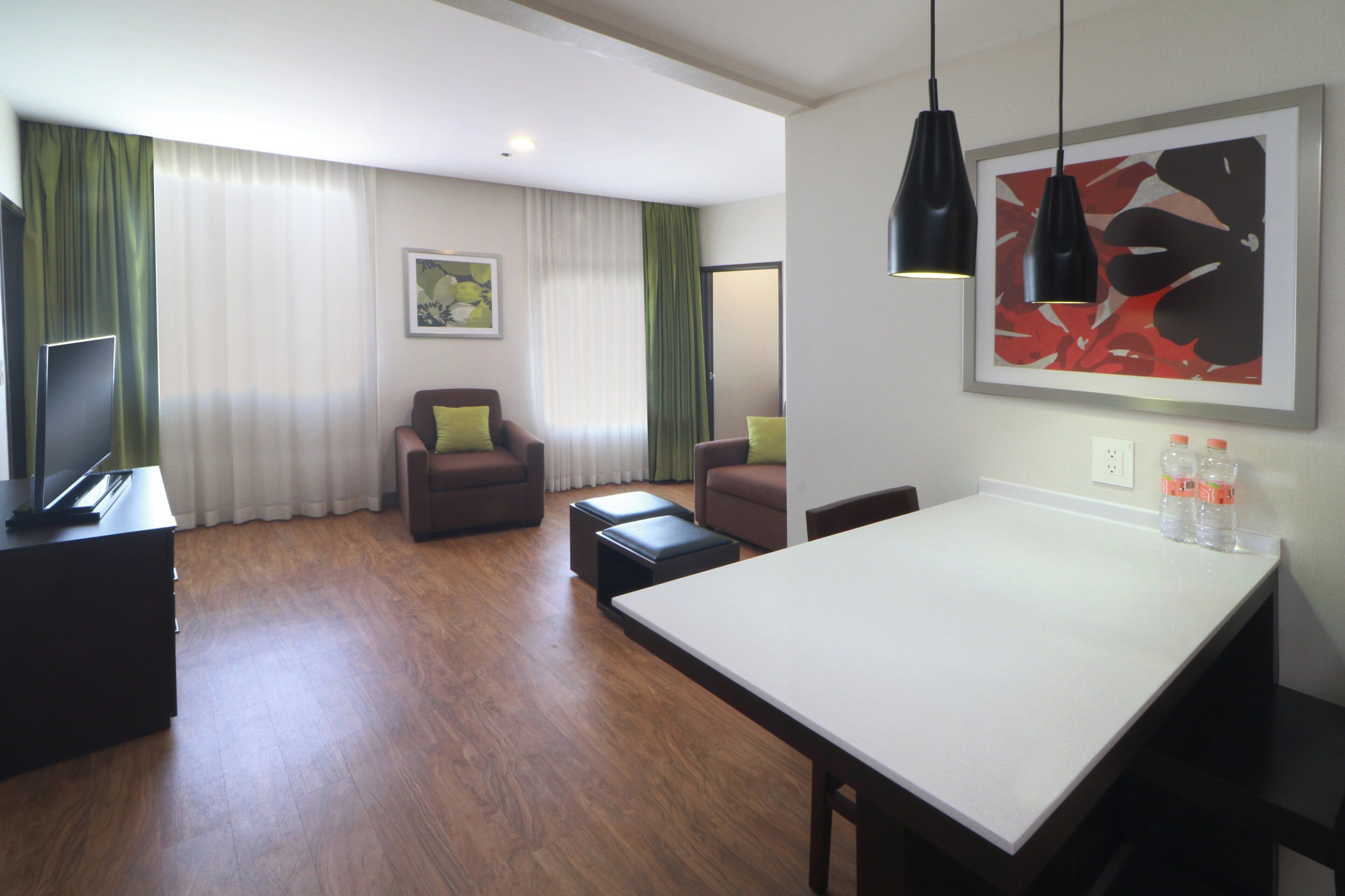 Una habitación de hotel moderna con zona de comedor, sofá, televisión y un cuadro en la pared.