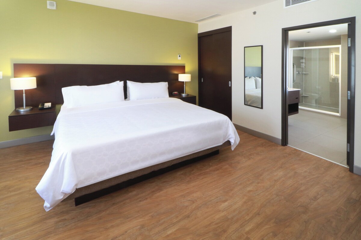 A hotel room with a large bed, two bedside tables with lamps, a mirror, and a door leading to a bathroom.