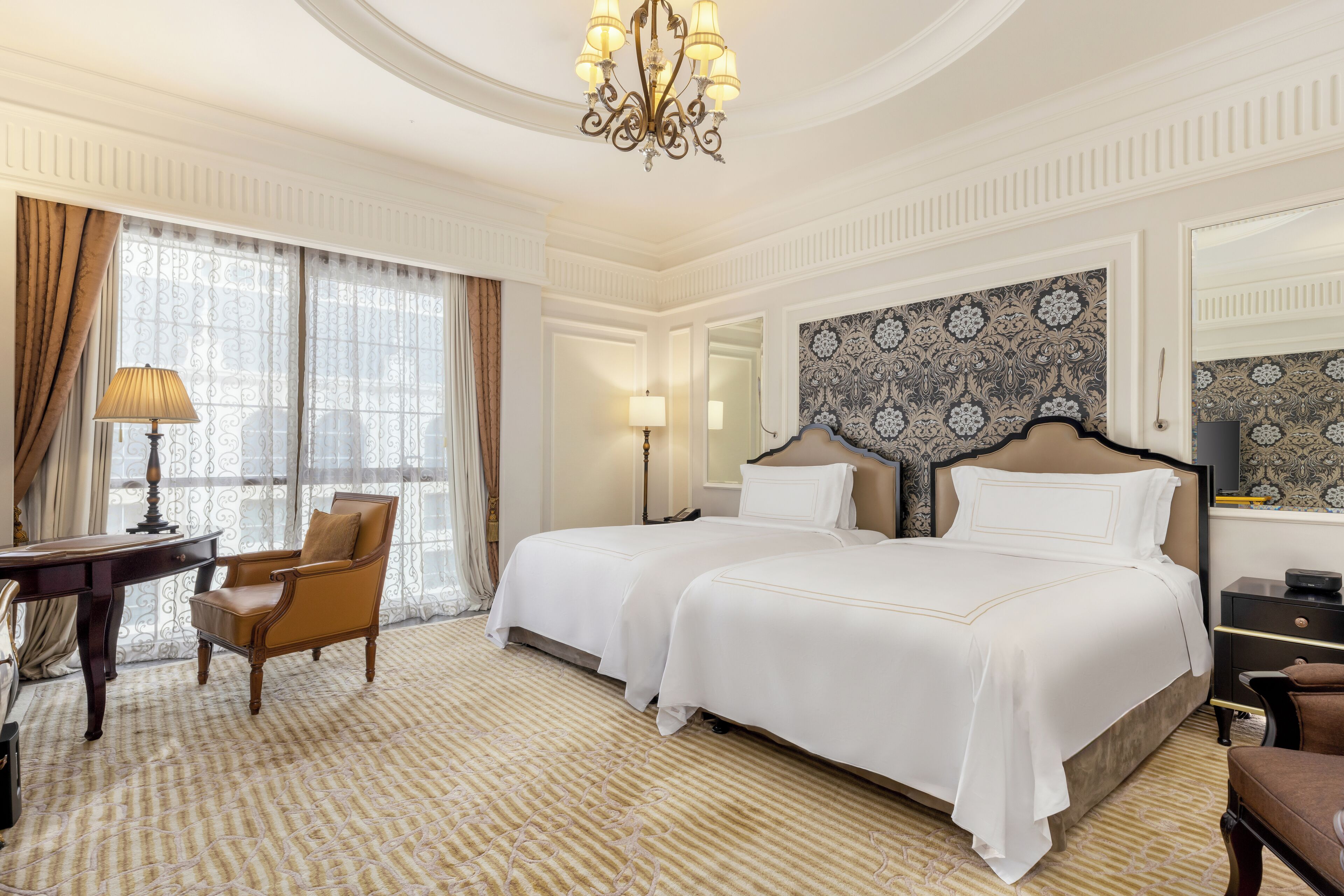 A hotel room with two beds, a desk, a chair, a chandelier, and patterned wallpaper.