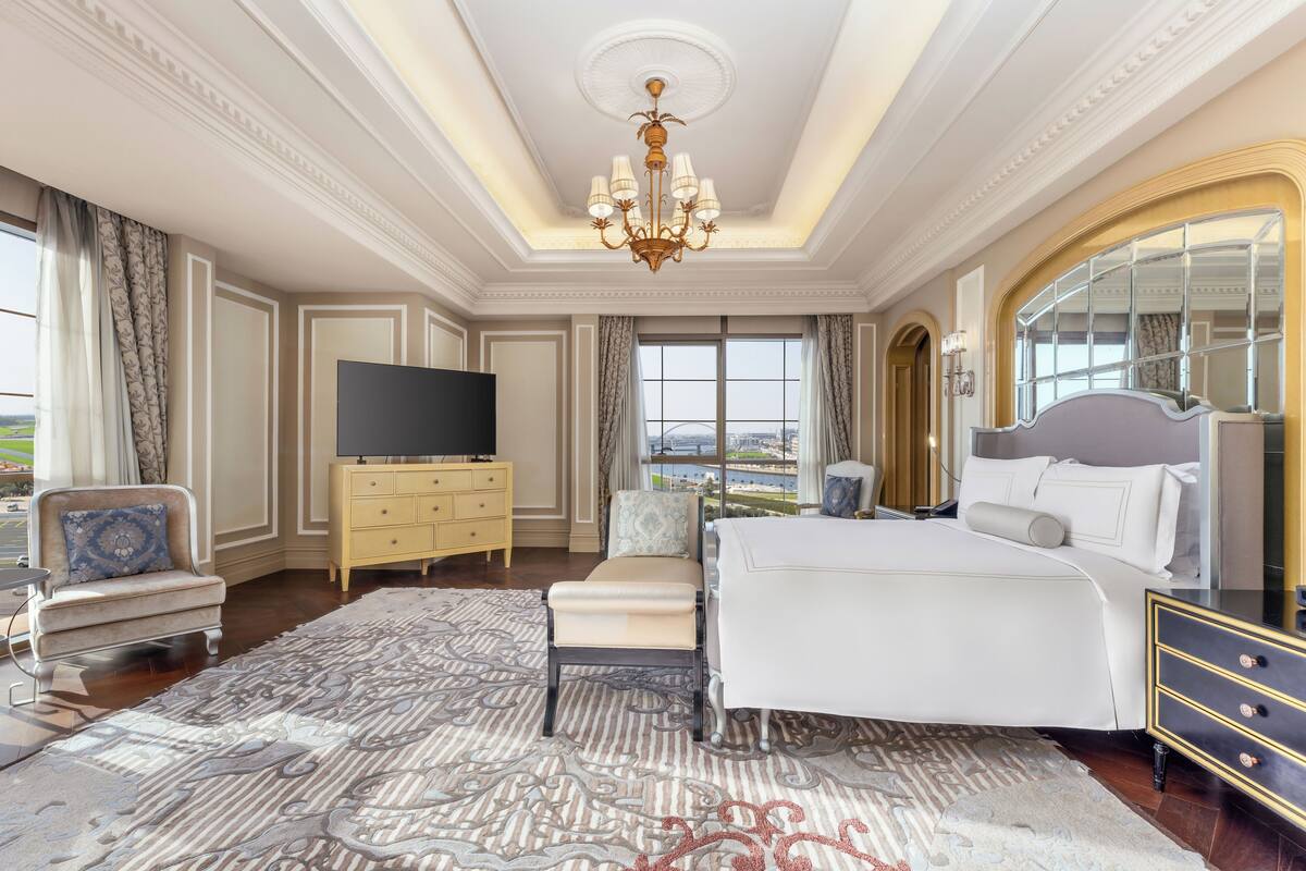 A spacious bedroom with a large bed, a television, and a view of the cityscape through the window.