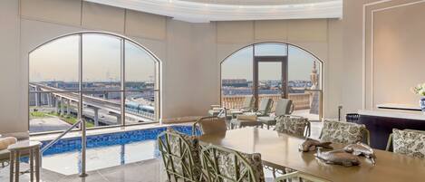 A spacious dining area with a large table and chairs, large windows offering a city view, and a pool area visible through the windows.