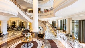 A grand lobby with a marble floor, a central round table with floral arrangements, and a grand staircase with ornate railings.