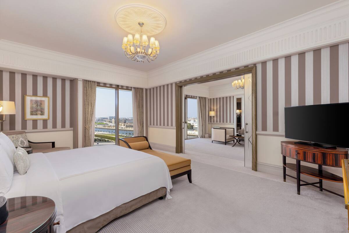A hotel room with a large bed, a television, a desk, and a view of the city through the windows.
