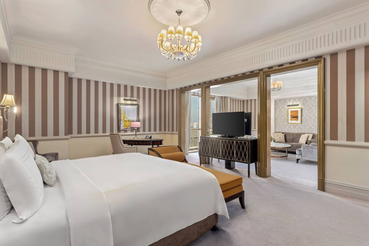 A hotel room with a large bed, a desk with a lamp, a television, and a view of the living area through an open door.