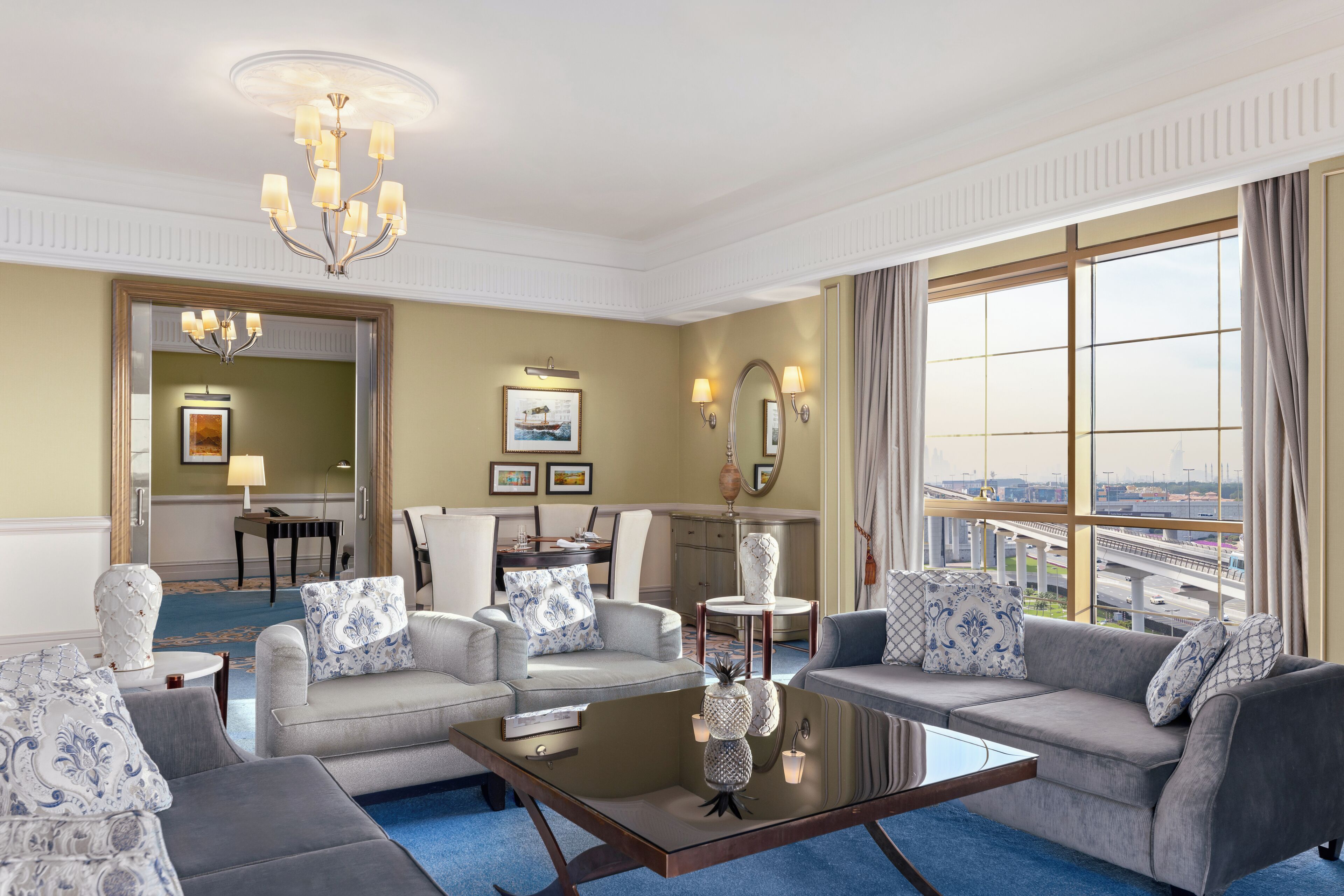 A spacious hotel room with a large window offering a city view, a chandelier, and a modern seating area with a coffee table.