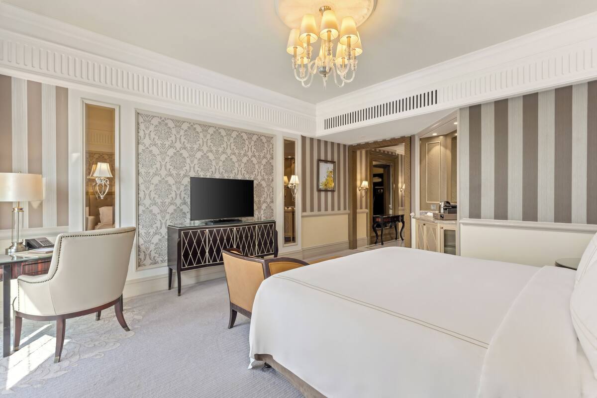 A hotel room with a large bed, a desk with a TV, two chairs, and a minibar.