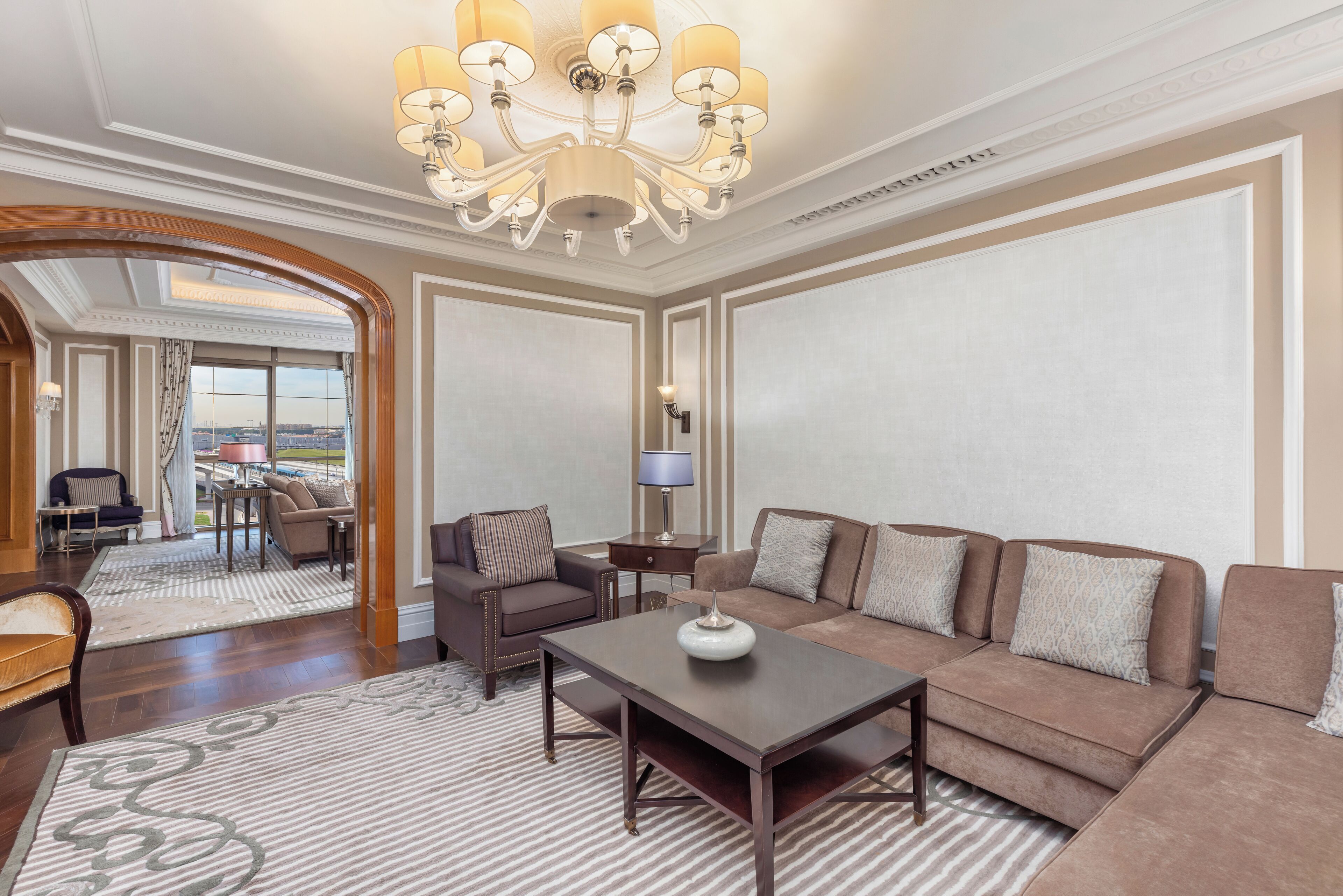 A spacious living room with a large sofa, a coffee table, and a chandelier. There is a view of the city through a large window.