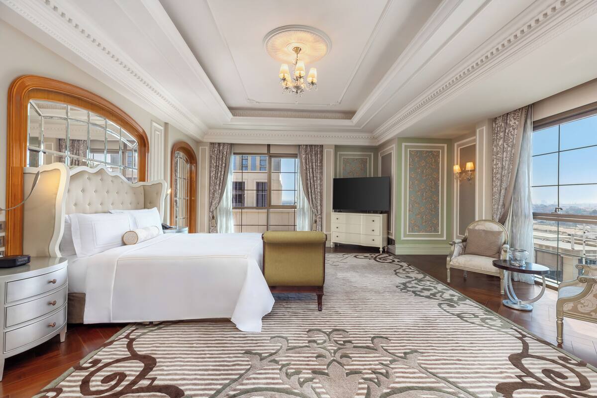 A luxurious hotel room with a large bed, a sitting area with a sofa and armchairs, a television, and a view of the city.