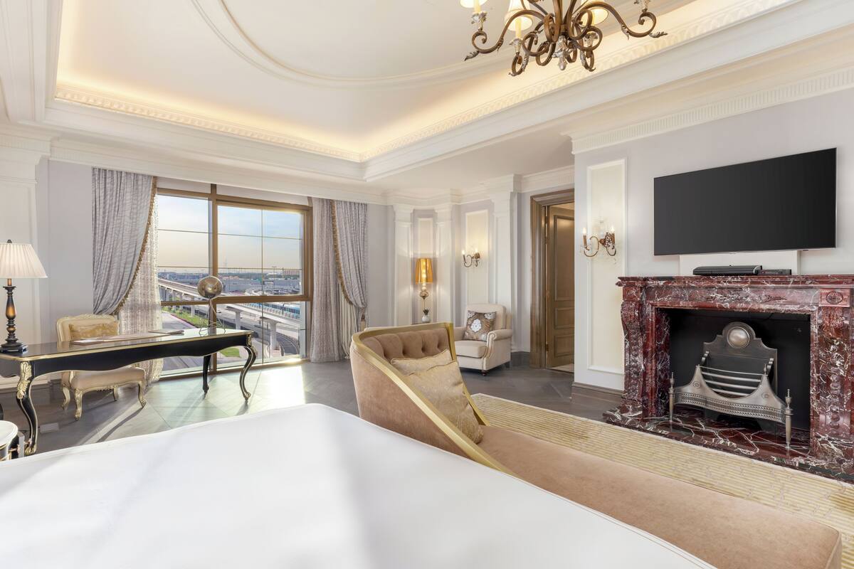 A luxurious hotel room with a large bed, a sitting area with a sofa and armchair, a fireplace, a television mounted on the wall, and a view of the city through large windows.