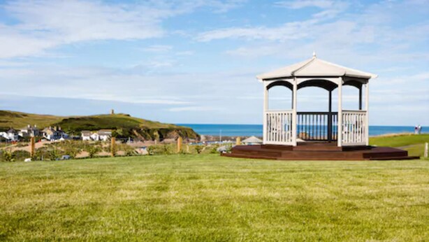 A gazebo with a wooden deck and railings, located on a grassy field with a view of the sea and houses in the background. - An Mor Hotel (Bude)