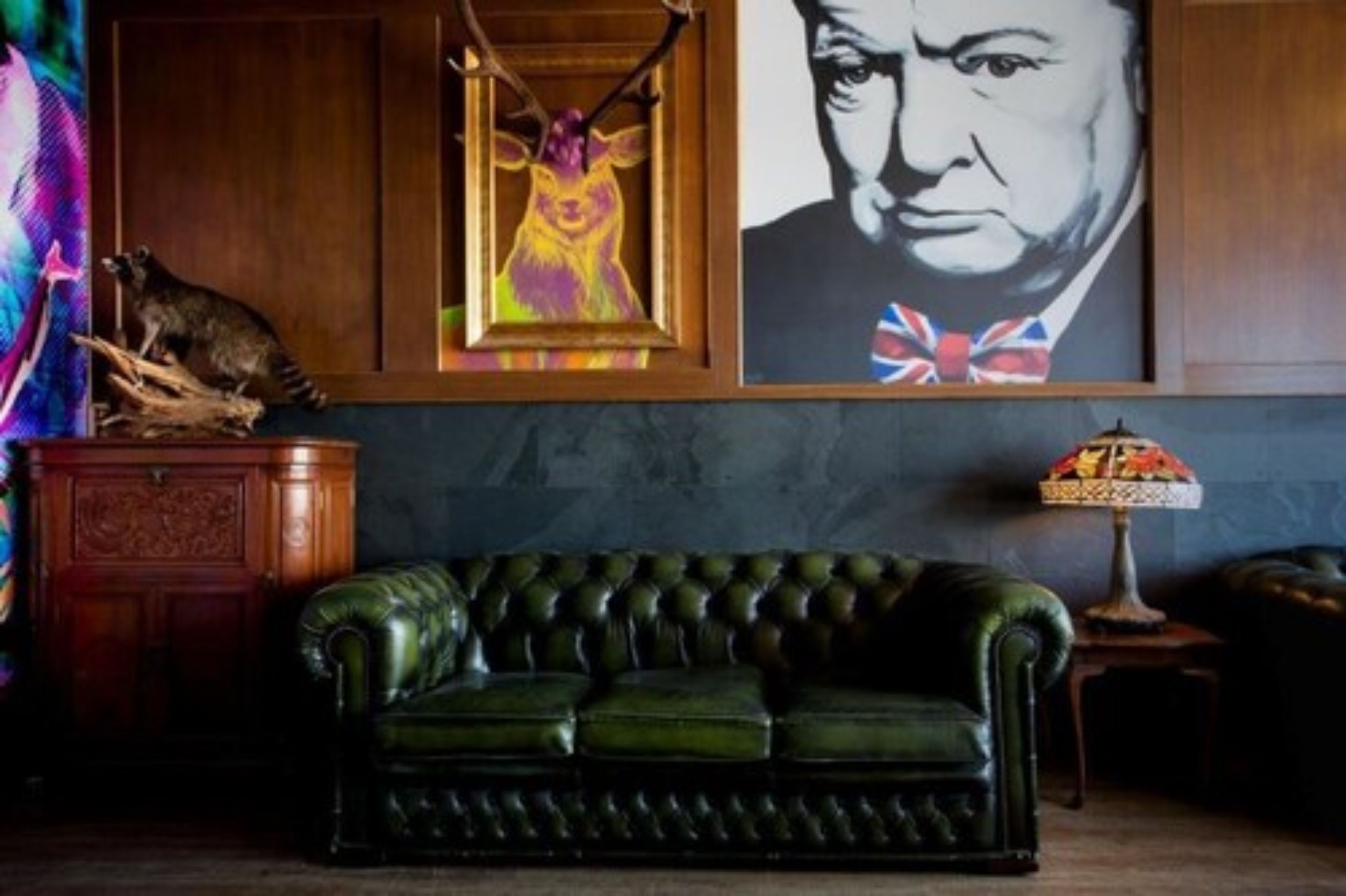 A green leather sofa in a room with a wooden cabinet, a framed painting of a deer, and a lamp on a side table.