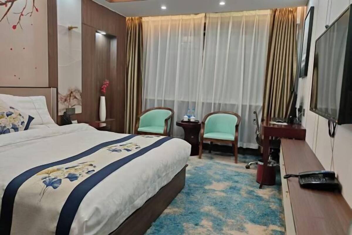 A hotel room with a bed, two chairs, a desk, a TV, and a window with curtains.
