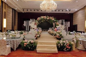 A stage with a floral arch, surrounded by tables and chairs, in a room with a chandelier. - Golmud Hotel (Golmud)