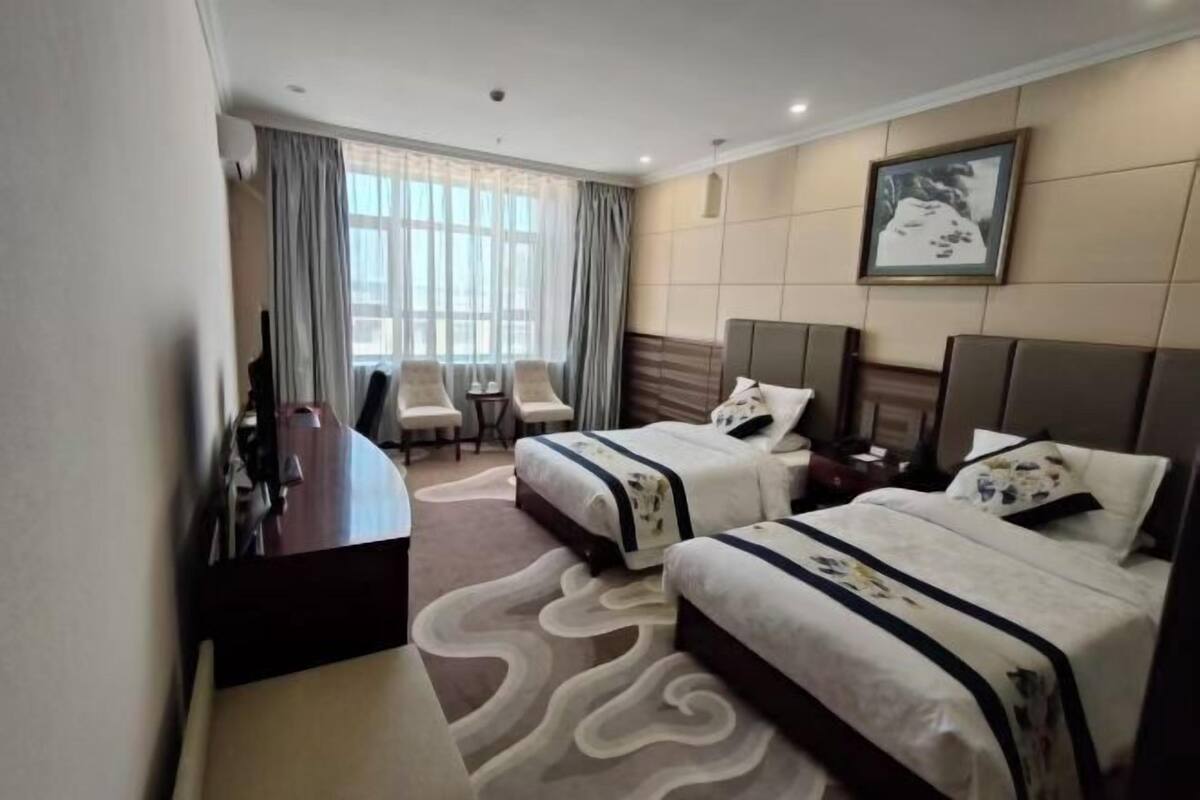 A hotel room with two beds, a desk, a chair, and a large window with curtains.