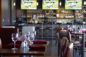 A restaurant with multiple tables, each set with wine glasses and napkins. There are televisions mounted on the wall displaying sports events. The bar area in the background has shelves stocked with bottles. - Four Points By Sheraton Midland (Midland)