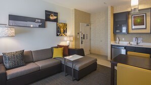 A modern hotel room with a sofa, ottoman, and a small dining table with chairs. There is a kitchen area with a dishwasher and a sink.
