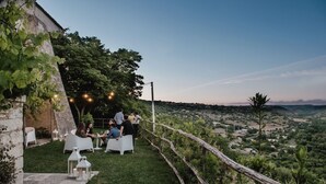An outdoor dining area with white tables and chairs, surrounded by greenery and overlooking a hilly landscape.