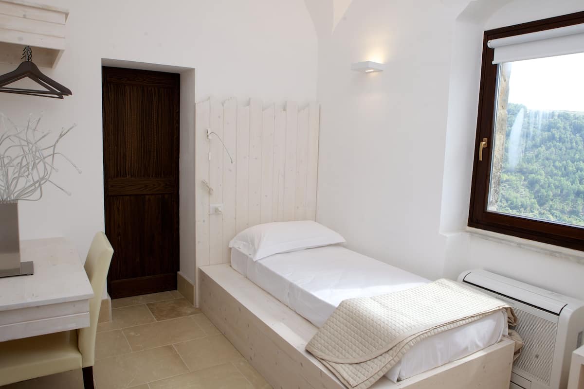 Una habitación pequeña y limpia con una cama, un escritorio y una ventana con vista a un bosque.
