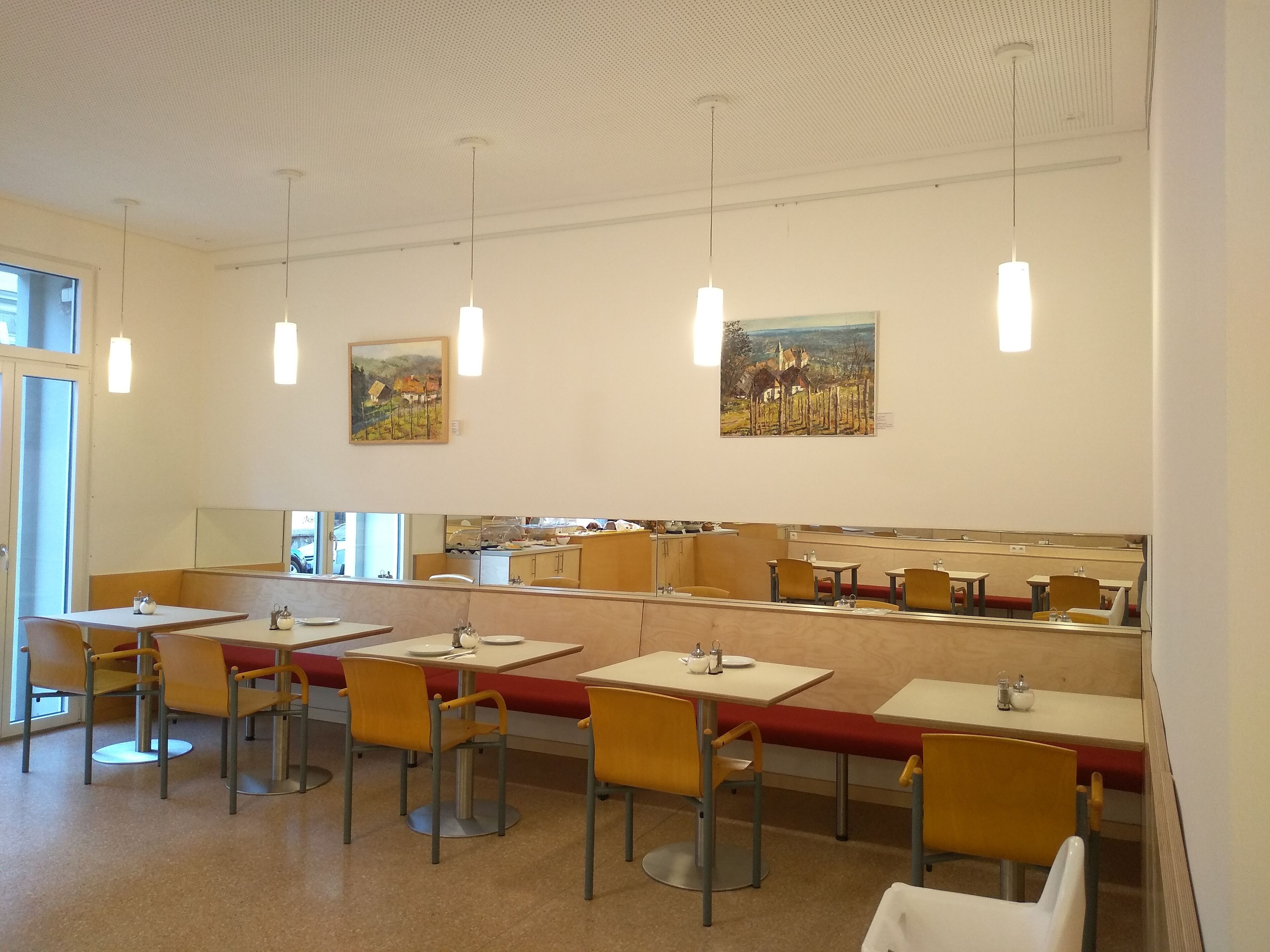 A dining area with tables and chairs, artwork on the walls, and a kitchen area in the background.