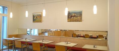 A dining area with tables and chairs, artwork on the walls, and a kitchen area in the background.