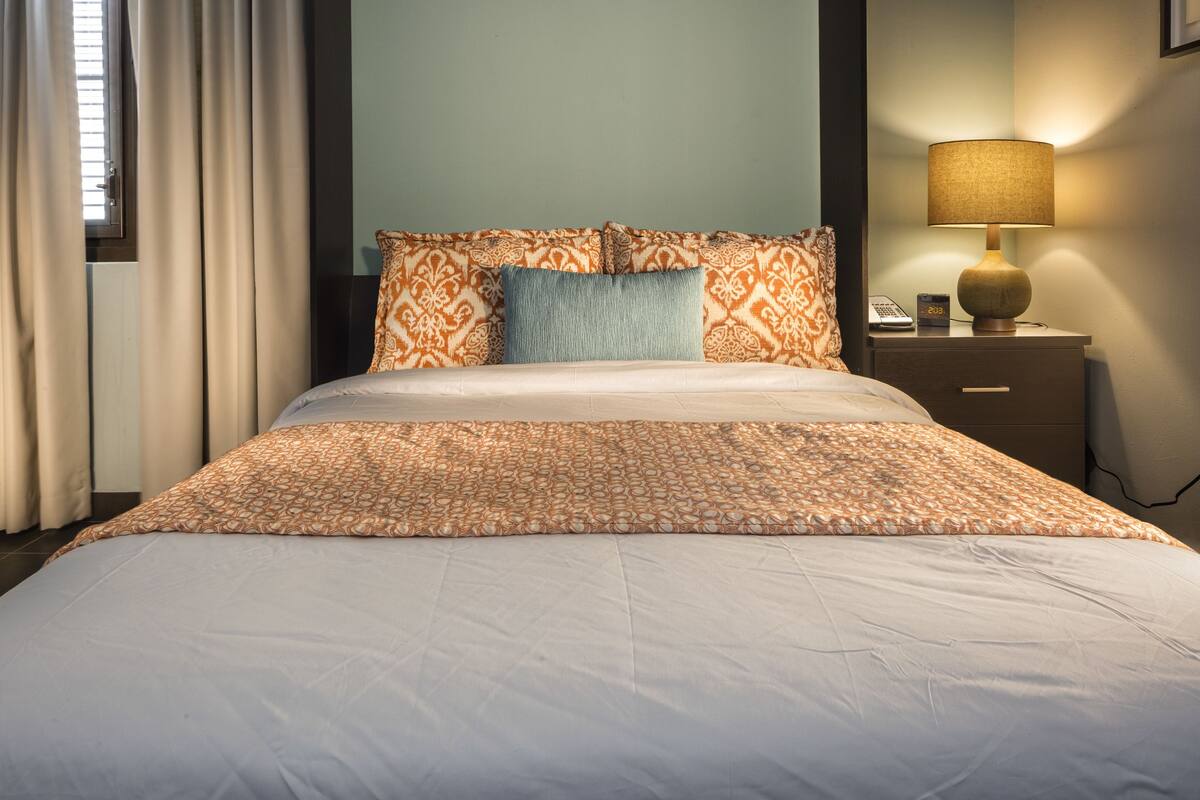 A neatly made bed with a patterned bedspread and pillows, a bedside lamp, and a nightstand with a phone and clock.