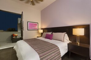 Una habitación de hotel con una cama, dos mesitas de noche, una lámpara de noche, una ventana y un ventilador de techo.