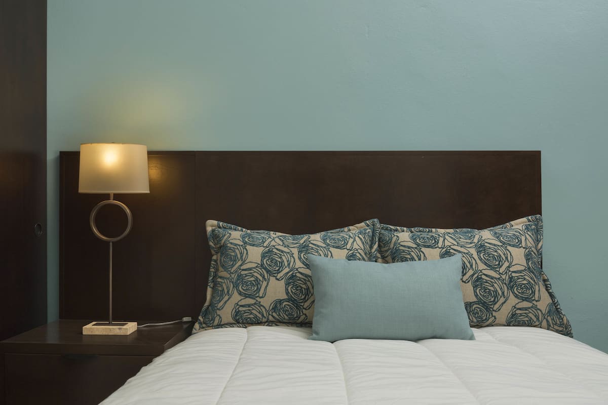 A neatly made bed with a white comforter and two pillows, one with a floral pattern and the other plain. A bedside table with a lamp and a power outlet is visible.