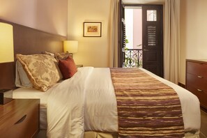 Habitación de hotel con una cama bien hecha, una mesita de noche con lámpara, una mesita auxiliar de madera y una puerta que da a un balcón.