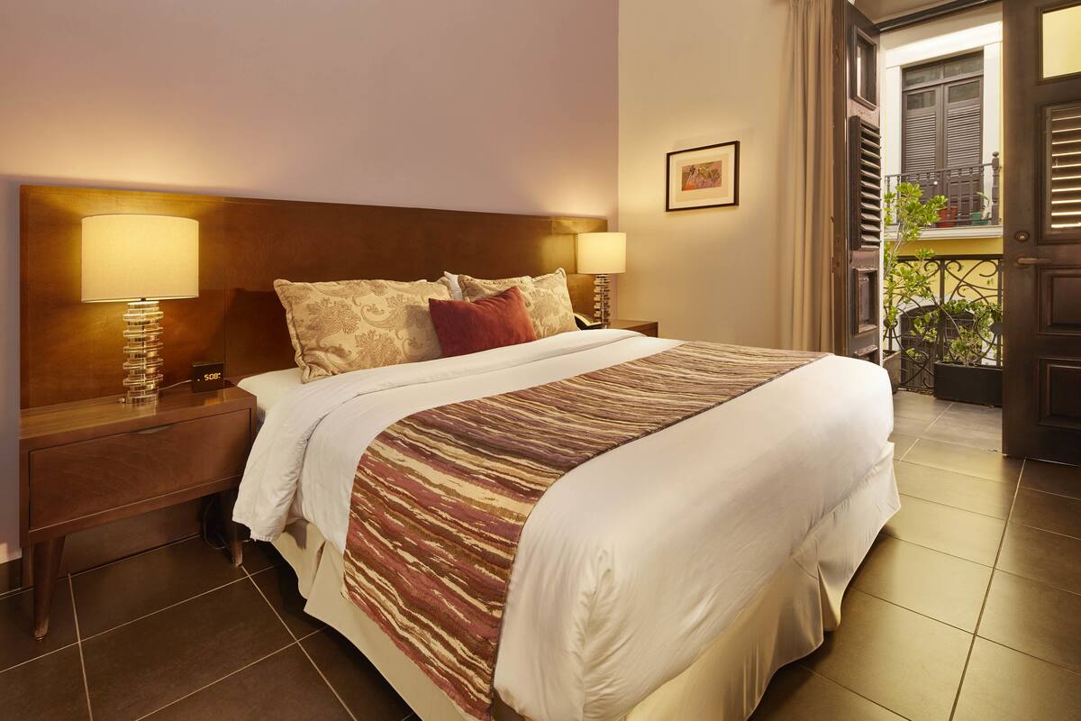 A neatly made bed with a patterned bedspread, two bedside lamps, a nightstand, and a view of the outside balcony.