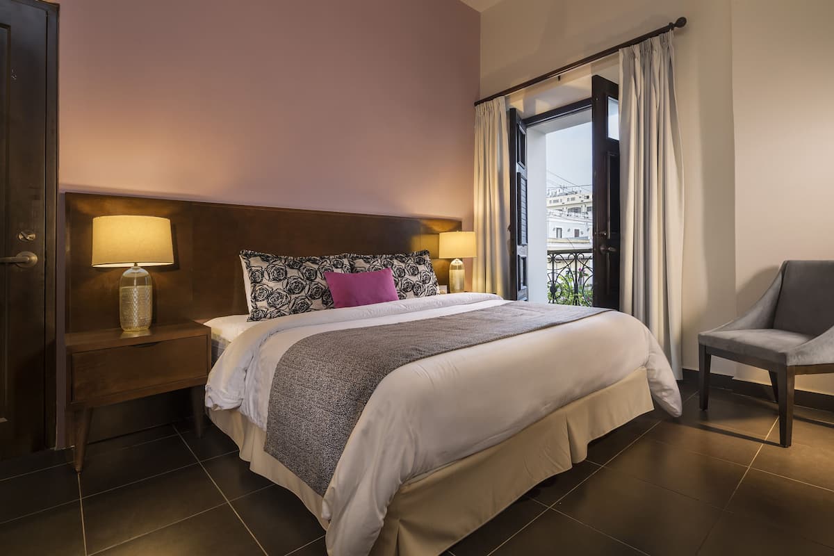 A hotel room with a large bed, two bedside lamps, a nightstand, a chair, and a view of the city through the window.