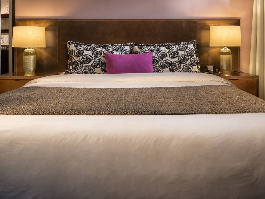 A neatly made bed with a patterned headboard, two bedside lamps, and a purple pillow.